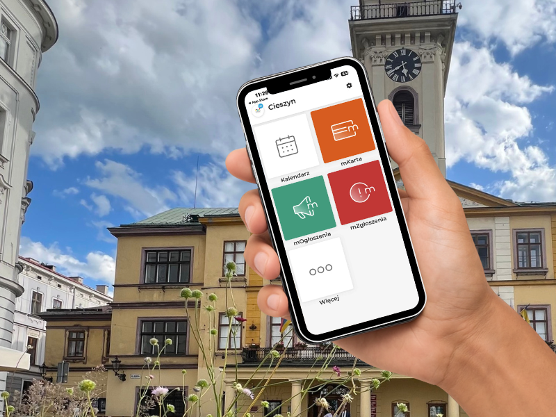 Na zdjęciu widać dłoń trzymającą smartfon na tle cieszyńskiego Ratusza. W telefonie jest otwarta aplikacja Cieszyn z kafelkami: Kalendarz, mKarta, Ogłoszenia, Zgłoszenia i Więcej. W tle znajduje się rynek z kamienicami oraz wieża z zegarem